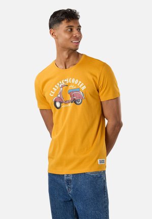 Mann trägt senfgelbes T-Shirt mit rotem Vintage-Roller-Motiv und blauen Jeans, schaut nach rechts, Hände hinter dem Rücken.