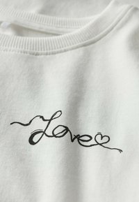 Sweatshirt blanc avec une finition texturée, arborant un design en cursive noire "Love" avec un accent en forme de cœur sur le devant.