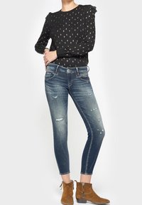 Top negro de manga larga con acentos estampados, combinado con jeans ajustados azules deslavados que presentan detalles desgastados y botines marrones.