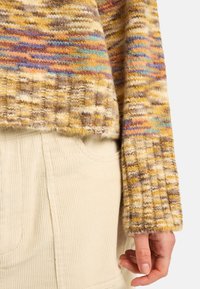 Wielokolorowy sweter z dzianiny z żółtymi, brązowymi i niebieskimi wzorami; ściągacze na rękawach; noszony na kremowym ubraniu z kordurowego materiału z widocznymi kieszeniami.