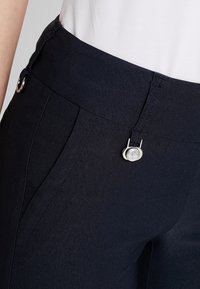 Nära bild av en person som bär skräddarsydda byxor i mörknavy med en silverfärgad rund metallspänne och en vit skjorta stoppad i.