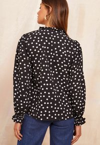 Blusa nera con pois bianchi, colletto arricciato e maniche gather. Indossata con pantaloni blu. Tessuto morbido, vestibilità comoda.
