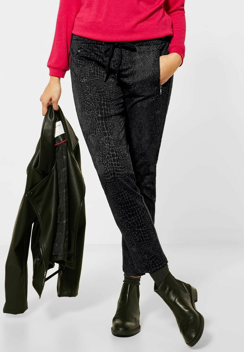 Personne portant un pantalon noir texturé, des bottes noires, un haut rouge à manches longues, tenant une veste en cuir noire dans une main.