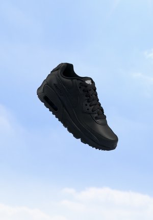 Chaussure de sport noire Nike avec lacets et semelle à air visible, montrée sur un fond de ciel bleu.