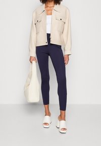 Béžová zkrácená bunda se dvěma předními kapsami, bílý top, námořnické legíny, bílé chunky sandály na podpatku a krémová kabelka přes rameno.