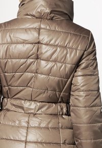 Brązowy ocieplany płaszcz z wysokim kołnierzem, z paskiem w talii i pikowanym materiałem. Tkanina ma błyszczące wykończenie i poziome szwy.