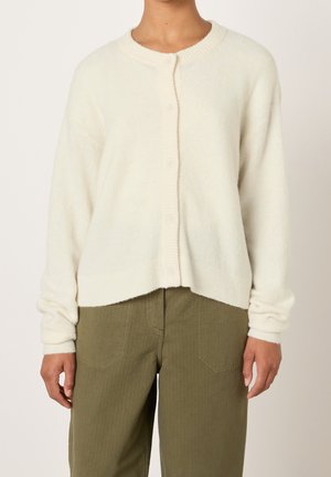 Vrouw draagt een zachte, crèmekleurige cardigan met knopen en geribbelde manchetten, gecombineerd met olijfgroene, hooggerichte broek met opgestikte zakken aan de voorkant.