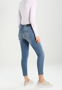 Tajta jeans i ljusblå denim med hög midja, två bakfickor och utskärningar vid ankelslitsen. Bärs med en vit topp och sneakers.