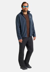 Mann in blauer Fleecejacke, braunem Hemd, schwarzen Hosen, schwarzen Schuhen und reflektierenden blauen Sonnenbrillen, der vor einem weißen Hintergrund steht.