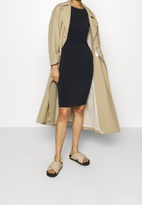 Beige trenchcoat över en figurnära marinblå klänning, tillsammans med tanfärgade slidesandaler. Släta texturer, kn älång klänning och ljusa accenter på trenchcoaten.