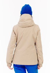 Beige waterdichte jas met een capuchon, elastische manchetten en verstelbare zoom. G gedragen over blauwe broek, met een soepele textuur en eenvoudig ontwerp.