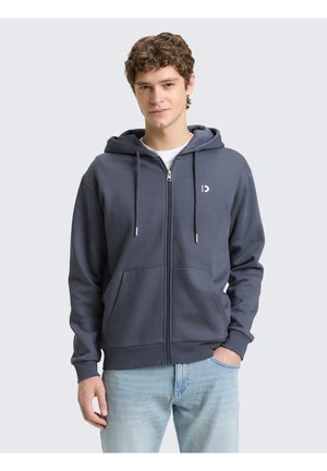 Junger Mann, der einen dunkelgrauen Kapuzenpullover mit Reißverschluss und Fronttaschen sowie hellblaue Jeans trägt, vor einem schlichten hellen Hintergrund steht.