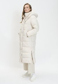 Manteau long matelassé de couleur beige clair avec capuche, poches latérales et fermetures à pression. Présente un design matelassé et un col montant pour plus de chaleur.