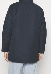 Laivastonsininen toppatakki, jossa korkea kaulus, pieni kolmivärinen logo selässä ja kyynärpääpaikkaukset. Kengurun taskut.