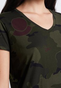 Grön camouflagemönstrad t-shirt med V-ringning, korta ärmar och en mjuk textur; har en vinröd cirkulär detalj och logotyp nära fållen.