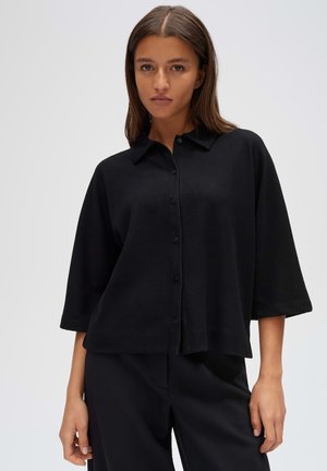 Donna con capelli castani lisci che indossa una camicia nera con bottoni e maniche larghe e pantaloni neri su uno sfondo chiaro e uniforme.
