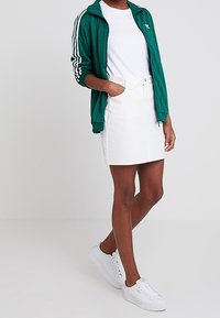 Grönrandig zip-up jacka med vita detaljer, kombinerad med en vit kjol. Modellen bär vita sneakers. Satt mot en neutral bakgrund.