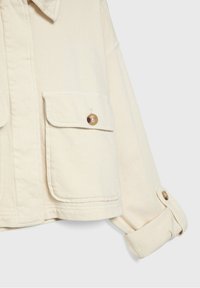 Manche de veste couleur crème avec poche à rabat boutonnée et poignet ajustable sur fond clair.