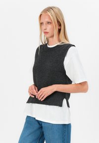 Pull en tricot sans manches de couleur charbon avec texture côtelée, col ras du cou, et fentes sur les côtés, porté sur un T-shirt blanc à manches courtes et un jean bleu.