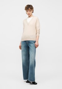 Personne portant un pull beige à col en V sur un haut blanc, un jean large bleu et des chaussures noires, debout devant un fond blanc.