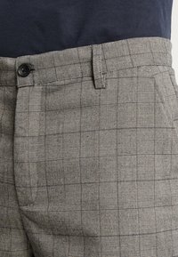 Pantalons à carreaux gris en tissu texturé, avec des poches latérales et un seul bouton noir à la taille. Détails cousus le long des coutures.