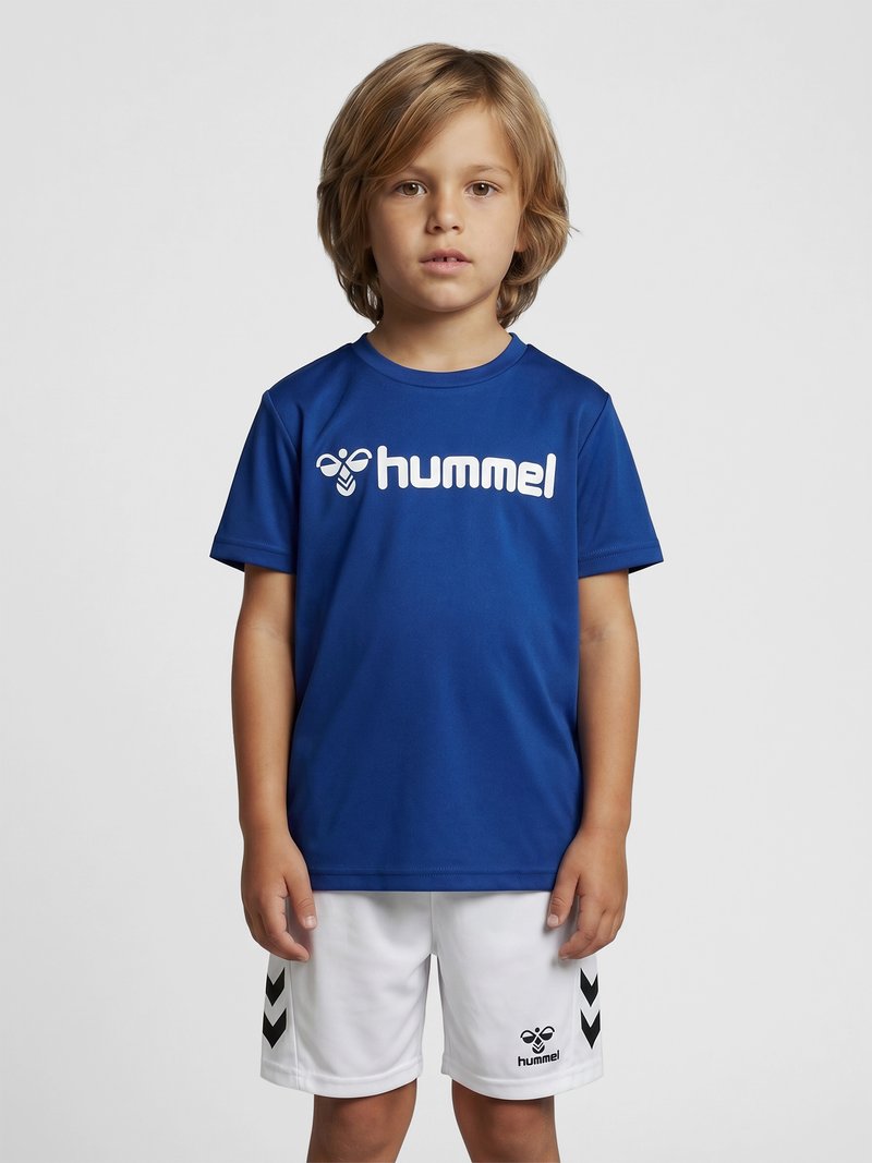 Jeune garçon debout portant un maillot de sport bleu hummel et un short blanc avec des logos chevrons noirs de hummel sur un fond gris uni.