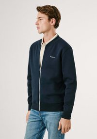 Mornarica zip-up dukserica s rebrastim ovratnikom i manžetama, s malim izvezenim logotipom na lijevoj strani grudi, u kombinaciji sa svijetloplavim trapericama.