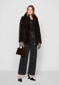 Veste en fausse fourrure marron, fermeture éclair, associée à un jean large noir déchiré, des gants en tricot gris et un petit sac à main marron. Mules à talons noirs.