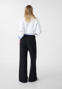 Weißer Pullover mit überschnittenen Schultern, cuffs in Blau und Weiß gestreift; kombiniert mit schwarzen, weit geschnittenen Hosen und weißem Schuhwerk. Sauberes, minimalistisches Design.