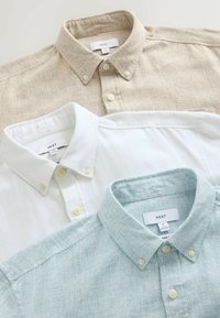 Trois chemises boutonnées en beige, blanc et bleu clair étalées à plat, chacune avec une étiquette de marque "NEXT" visible sur le col.