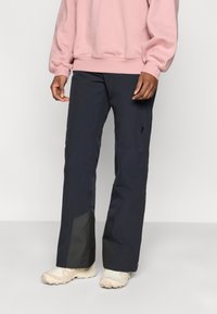 Αδιάβροχο παντελόνι σε ναυτικό μπλε με φαρδιά γραμμή, με μαύρο λεπτομέρεια στο τελείωμα, συνδυασμένο με ανοιχτό ροζ φούτερ και μπεζ αθλητικά παπούτσια.