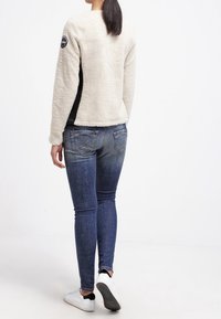 Kremowa kurtka z podszyciem z polaru, czarne akcenty, okrągły dekolt i naszywka z logo, zestawiona z dopasowanymi niebieskimi dżinsami i białymi trampkami.