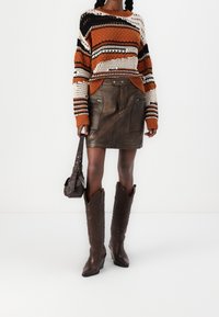 Neulottu neule, jossa oransseja, kermaisia ja mustia raitoja; ruskea nahkahame vetoketjuilla; ruskeat cowboy-saappaat koristeellisella kirjonnalla.