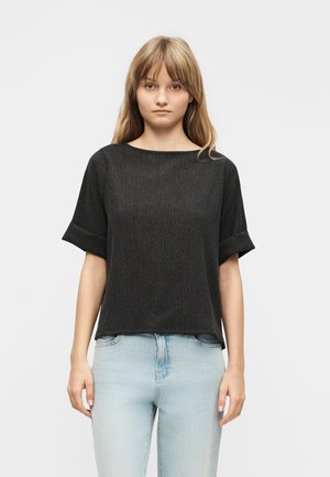 Jeune femme aux cheveux châtain clair raides, portant un haut noir à manches courtes texturé et un jean bleu clair, faisant face à l'avant avec une expression neutre.