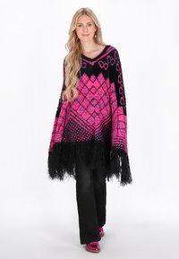 Poncho tricotat în roz și negru cu modele geometrice, tiv franjurat, design cu decolteu în v. Este asortat cu pantaloni negri și pantofi roz strălucitori.