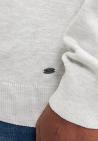 Maglione grigio chiaro in maglia, con polsini e fondo a costine, e un piccolo etichetta nera con il logo "Petrol" sul lato sinistro in basso.