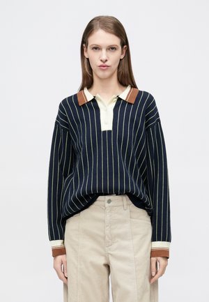 Pulover bleumarin cu dungi verticale ivoire, guler clasic în nuanță crem deschis și maro; mâneci largi, croială lejeră, manșete nervurate. Pantaloni bej din catifea.