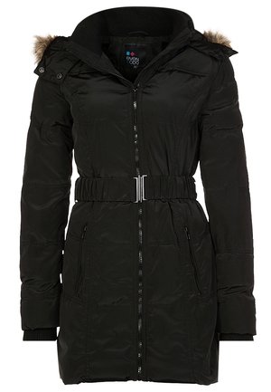 Femme portant un manteau d'hiver noir à capuche avec bordure en fourrure, un t-shirt gris foncé, un jean bleu et des baskets Adidas noir et blanc, debout avec une main dans la poche.