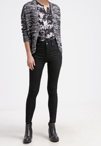 Kvinna iklädd svarta högmidjade skinny jeans, svart blommig blus, grå mönstrad öppen cardigan och svarta ankelstövlar framför en enkel vit bakgrund.