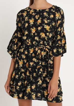 Femme portant une robe noire à motifs floraux avec des fleurs orange et jaunes, dotée de manches évasées et d'une ceinture à nouer à la taille.