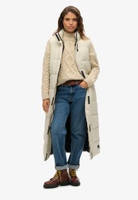 Gilet imbottito lungo beige con colletto alto e zip frontale, indossato sopra un maglione lavorato a maglia crema, jeans blu e stivali marroni con lacci.