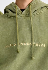 Olive spalvos džemperis su kapišonu, pagamintas iš minkštos medžiagos, su priekyje esančia kišene, užtrauktuku reguliuojamu kapišonu ir "ALPHA INDUSTRIES" siuvinėjimu smėlio spalvos.