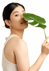 Una persona tiene in mano una grande foglia verde di Monstera, indossando una canotta beige chiaro. Lo sfondo è bianco, mettendo in risalto il soggetto e la foglia.