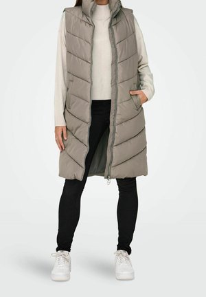 Person trägt eine lange, ärmellose beige Steppweste über einem cremefarbenen Pullover, schwarze Hose und weiße Turnschuhe und steht mit einer Hand in der Tasche.