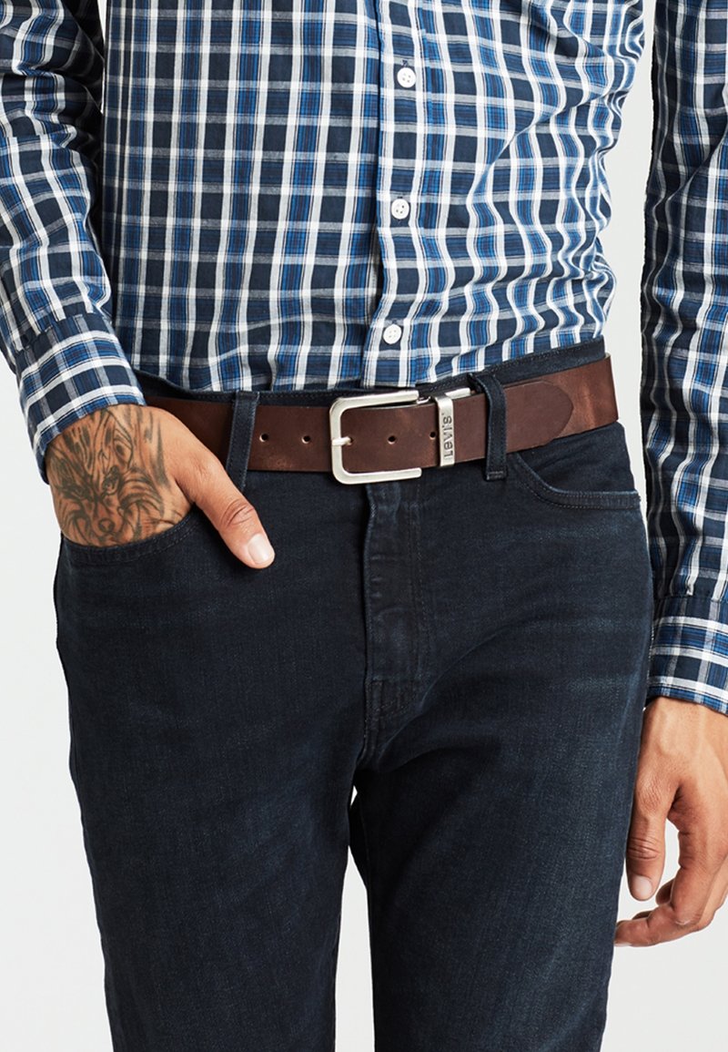 Homme portant une chemise à carreaux bleue et blanche rentrée dans un jean foncé avec une ceinture en cuir marron Levi's, main gauche dans la poche, tatouage visible sur le poignet.