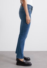 Jeans de mezclilla azul con campana y dobladillo deshilachado, ajustados en las caderas y los muslos, que presentan dos bolsillos traseros y una etiqueta de cuero marrón.