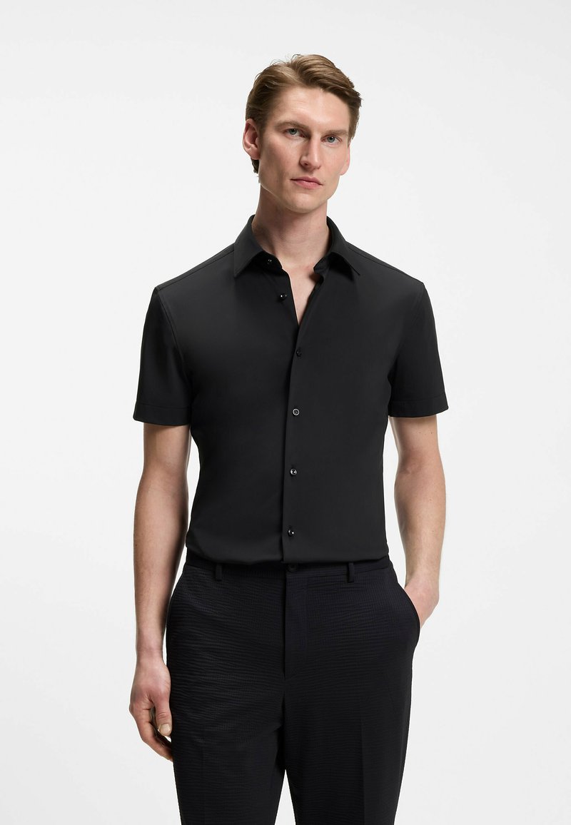 Homme aux cheveux châtain clair courts portant une chemise noire ajustée à manches courtes boutonnée et un pantalon noir sur un fond uni.