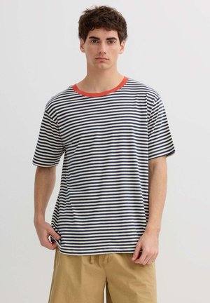 Joven con camiseta de rayas azul marino y blanco con cuello naranja y pantalones cortos beige, posando frente a un fondo claro y liso.