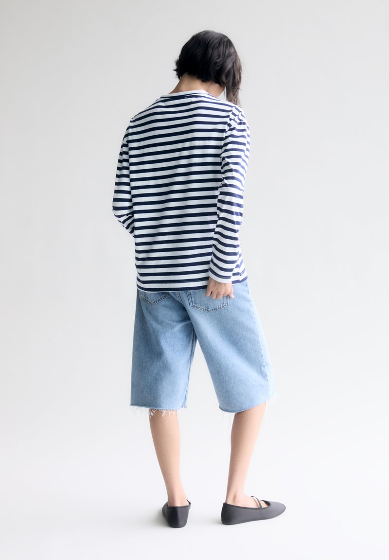 Camicia a maniche lunghe a righe blu navy e bianca, abbinata a pantaloni corti in denim azzurro chiaro. I pantaloni corti presentano bordi sfrangiati e una vestibilità rilassata.