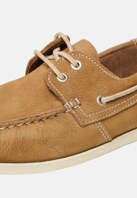 Hellbraunes Bootsschuh aus Leder mit strukturiertem Obermaterial, beigen Schnürsenkeln, silbernen Ösen und weißen Nähten entlang der Ränder.